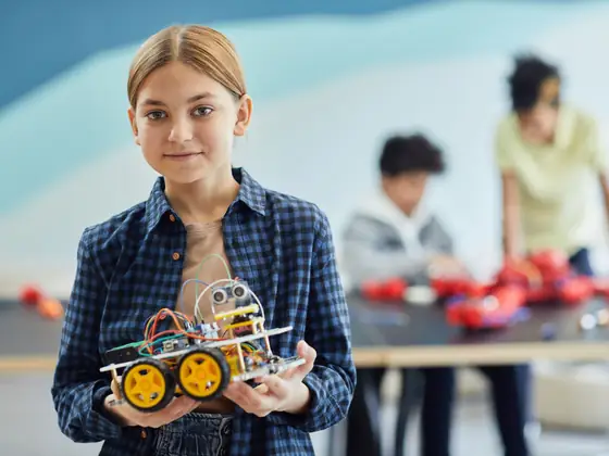 A girl holding the robot she built, grinning at the camera