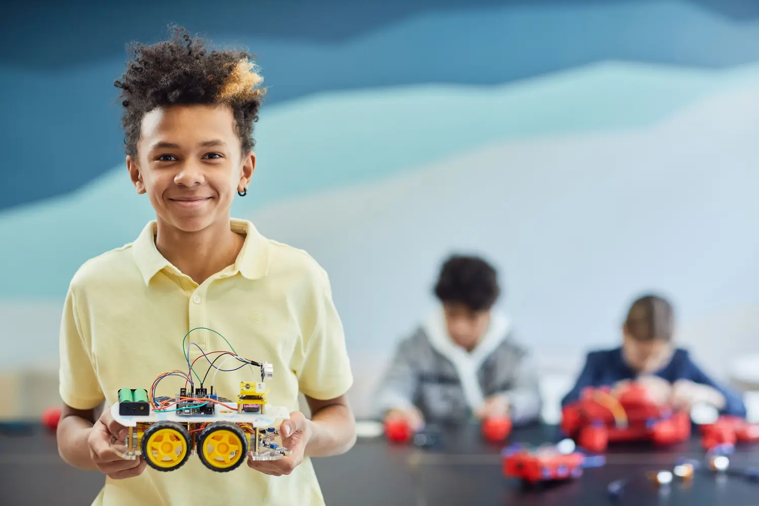 A smiling boy holding up the wheeled robot he built, with classmates working on their own robots behind him