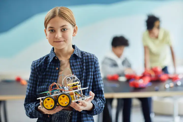 A girl smiling proudly as she holds up the robot she assembled in class